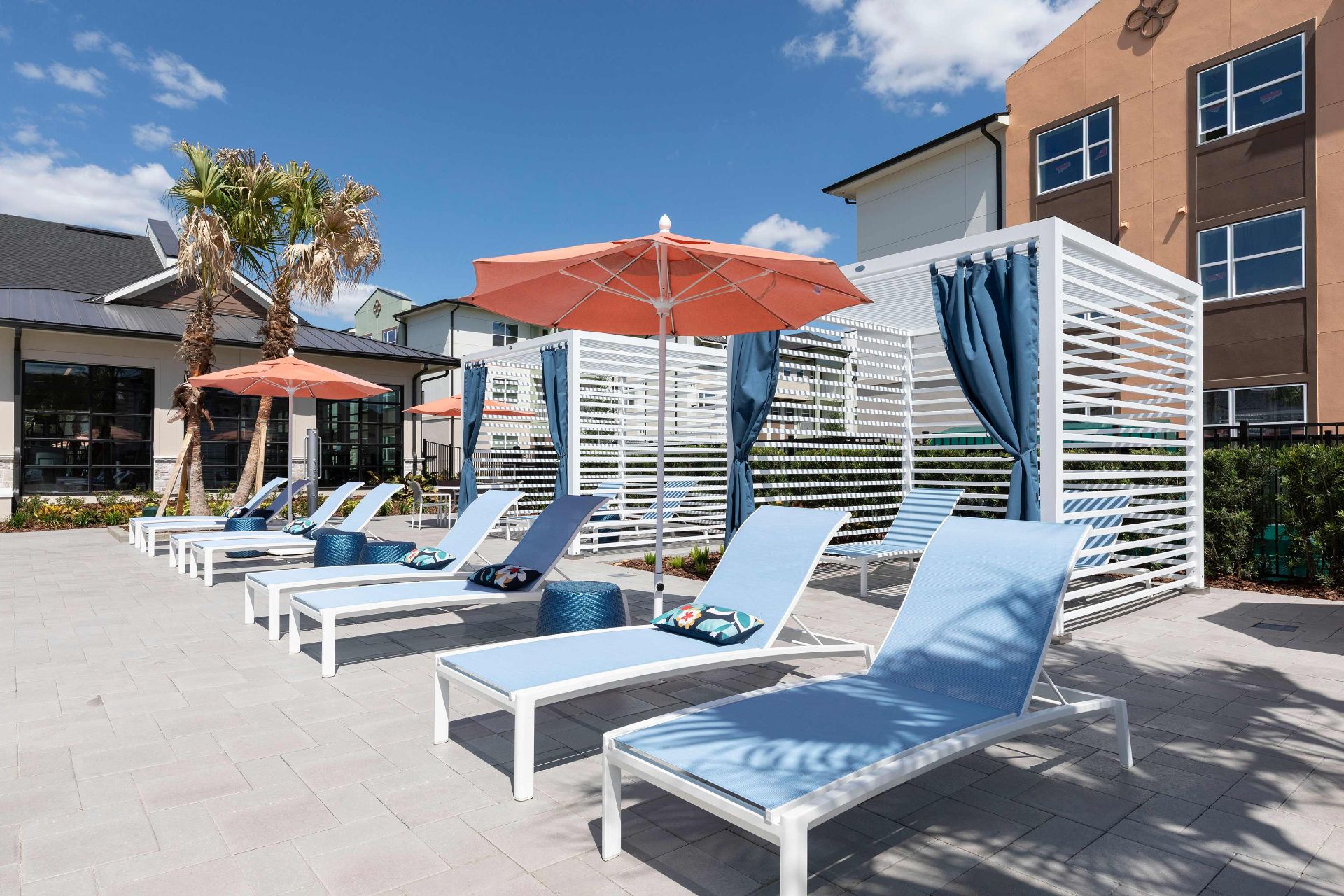 Poolside lounge area with blue sunbeds, orange umbrellas, and white cabanas with blue curtains, adjacent to a modern building under a sunny sky.