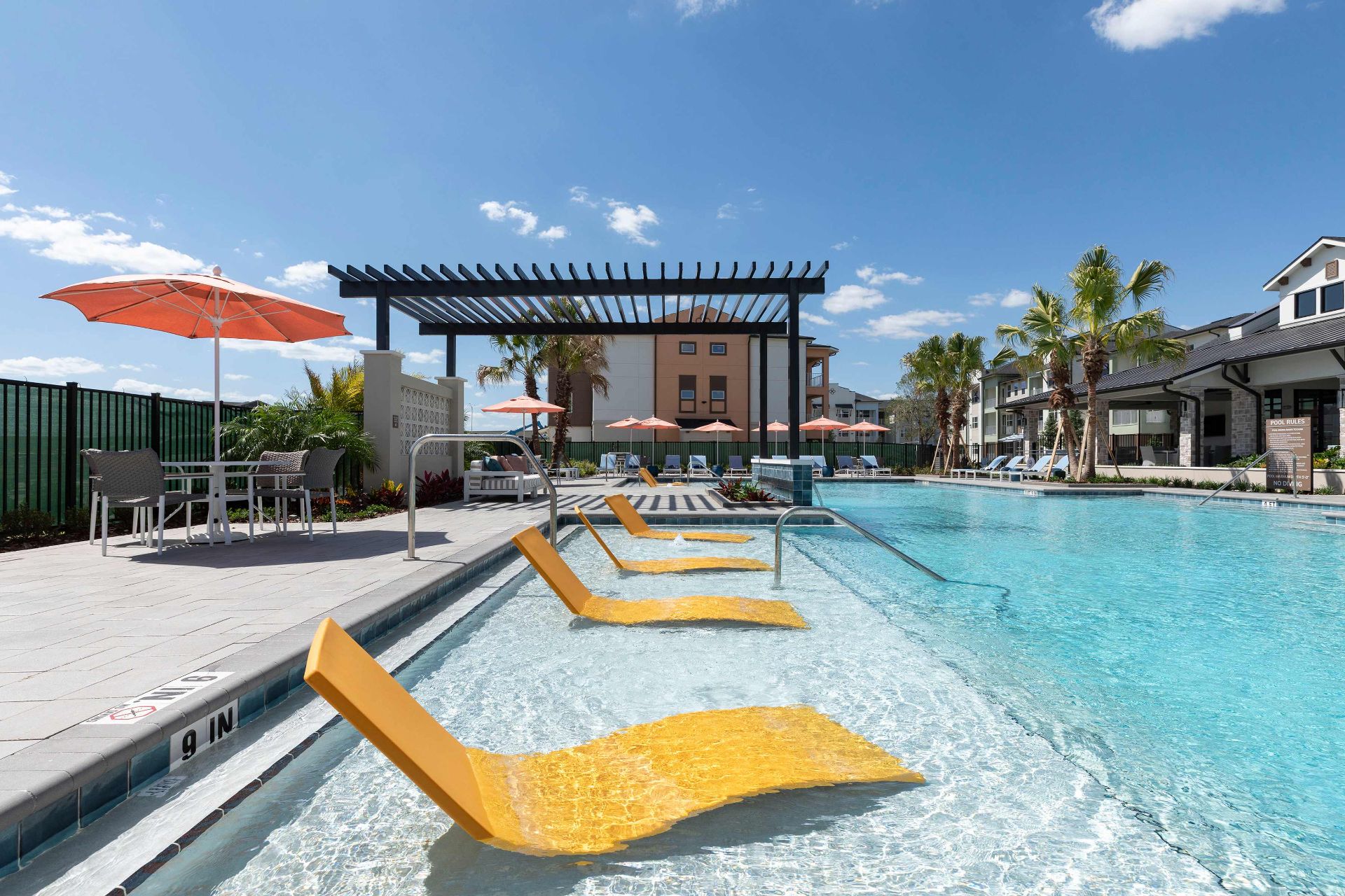 Outdoor swimming pool with yellow lounge chairs partially submerged in water, tables with orange umbrellas, palm trees, and apartment buildings in the background under a blue sky.
