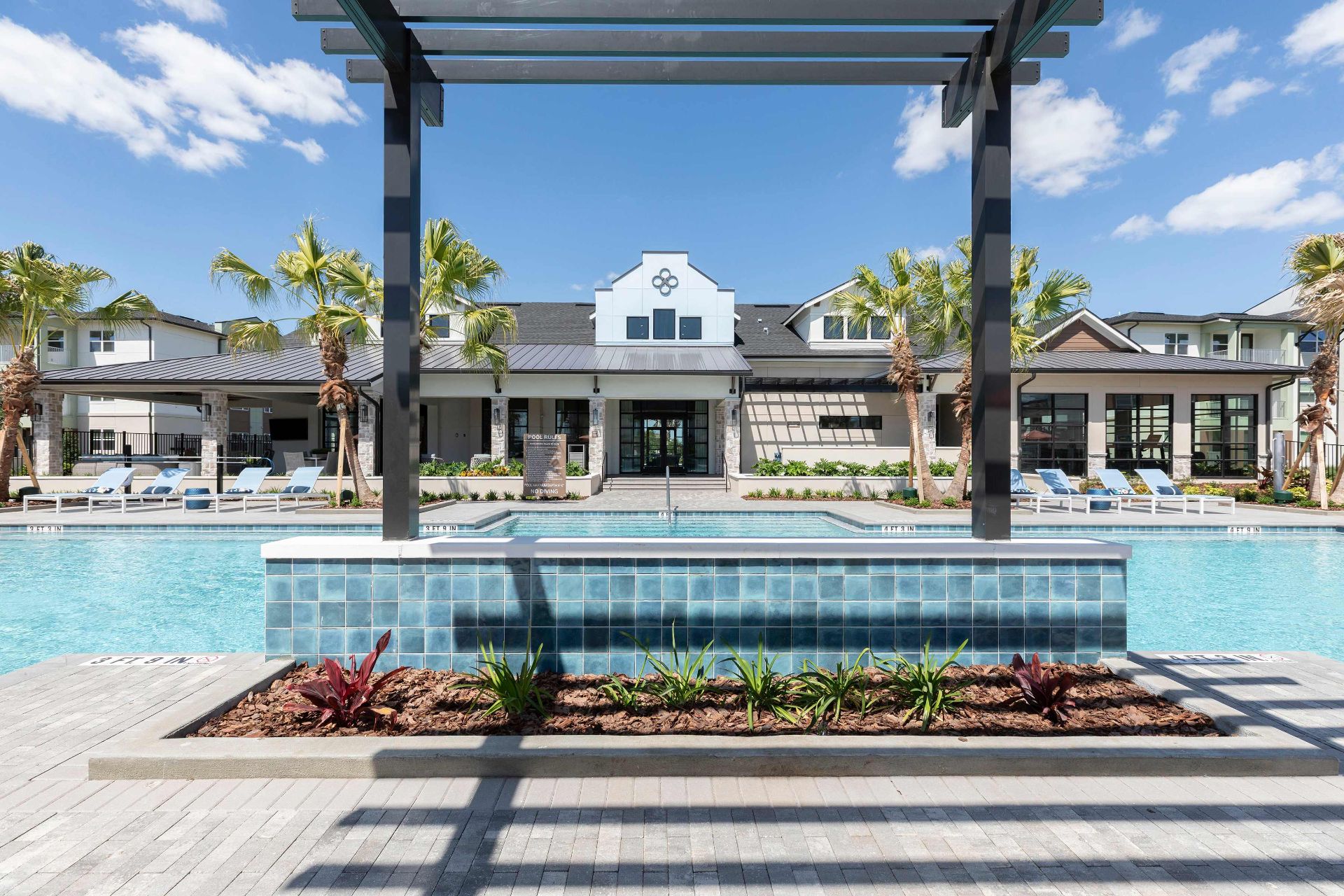 Outdoor swimming pool with lounge chairs, palm trees, and a modern clubhouse building in the background under a clear blue sky.