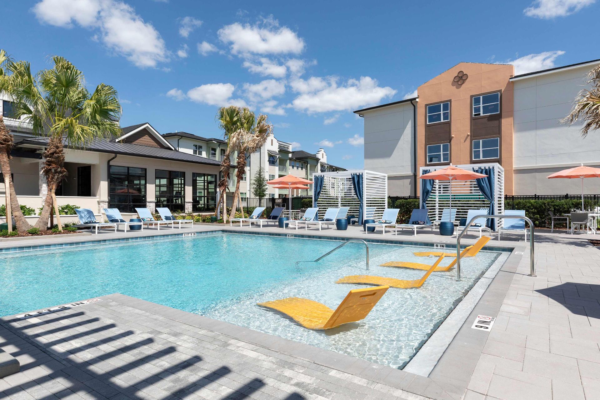 Outdoor swimming pool with yellow lounge chairs in the shallow end, surrounded by sunbeds, umbrellas, palm trees, and modern apartment buildings under a blue sky.