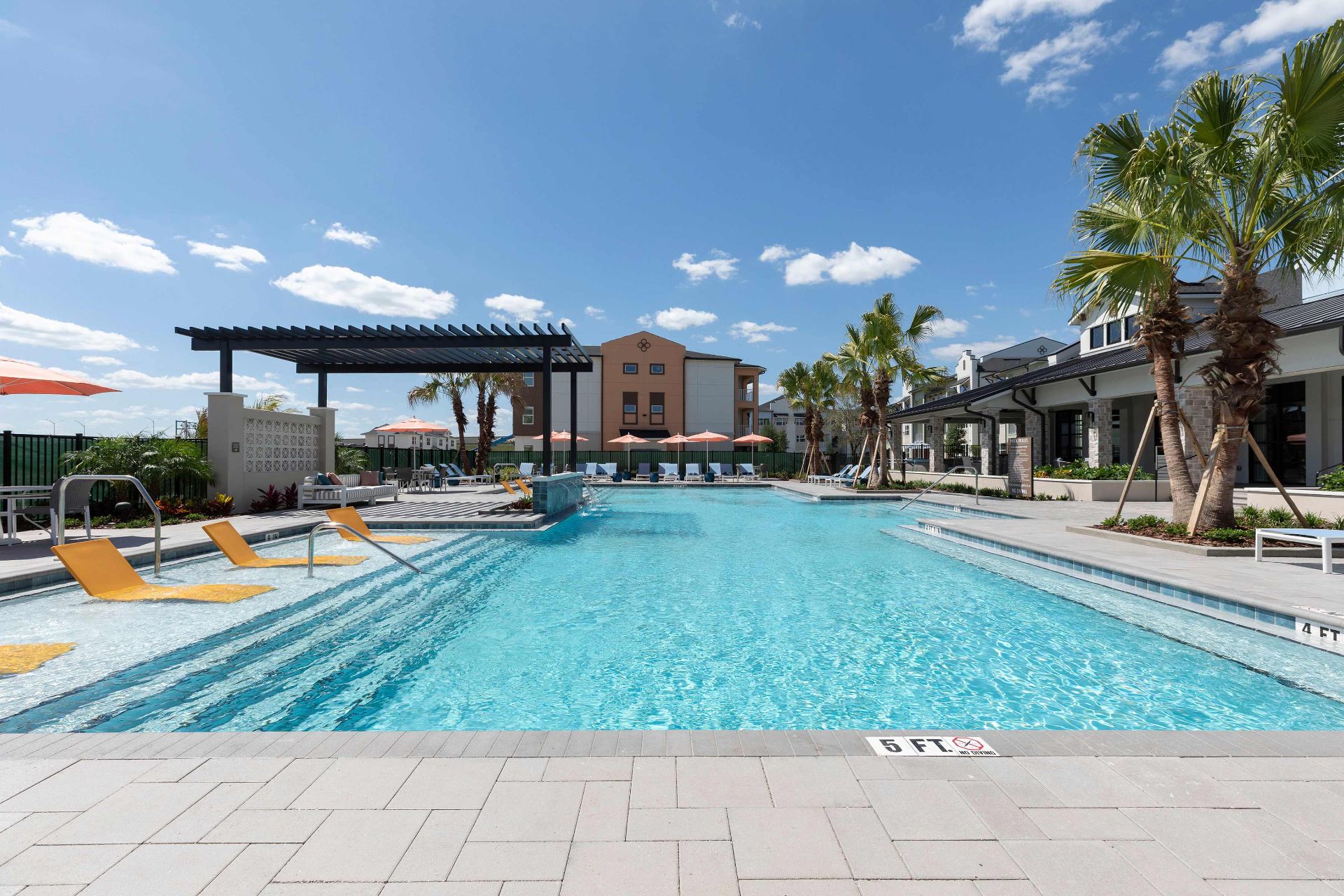 Outdoor swimming pool with lounge chairs, palm trees, shaded seating areas, and buildings in the background under a clear blue sky.