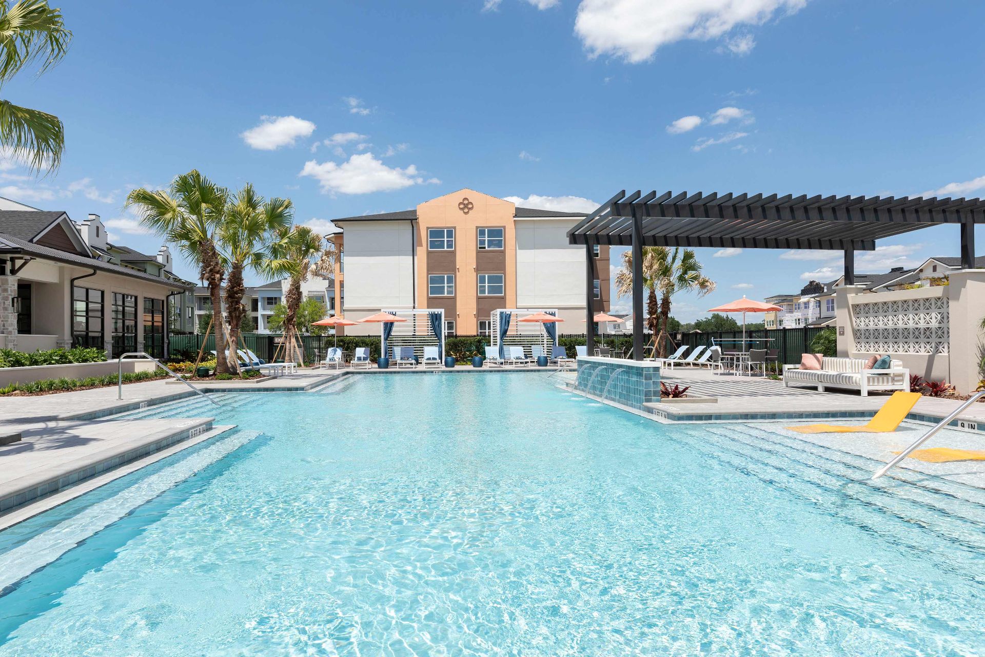 Outdoor swimming pool with lounge chairs, umbrellas, and shaded seating area in front of a modern apartment building on a sunny day.