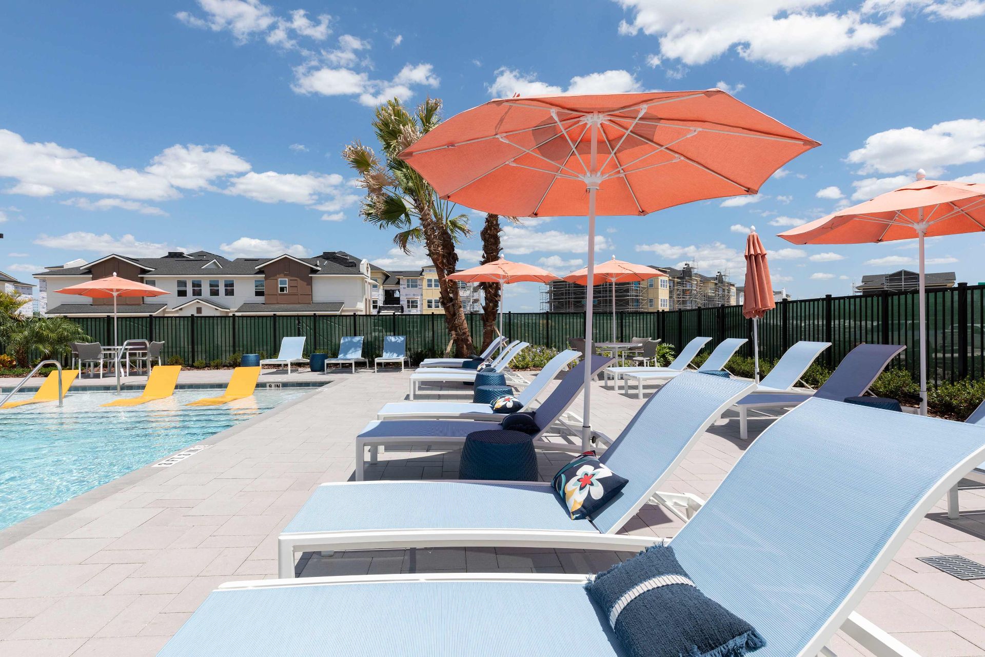 Poolside lounge chairs with blue cushions and orange umbrellas beside a clear swimming pool; residential buildings visible in the background under a blue sky with clouds.