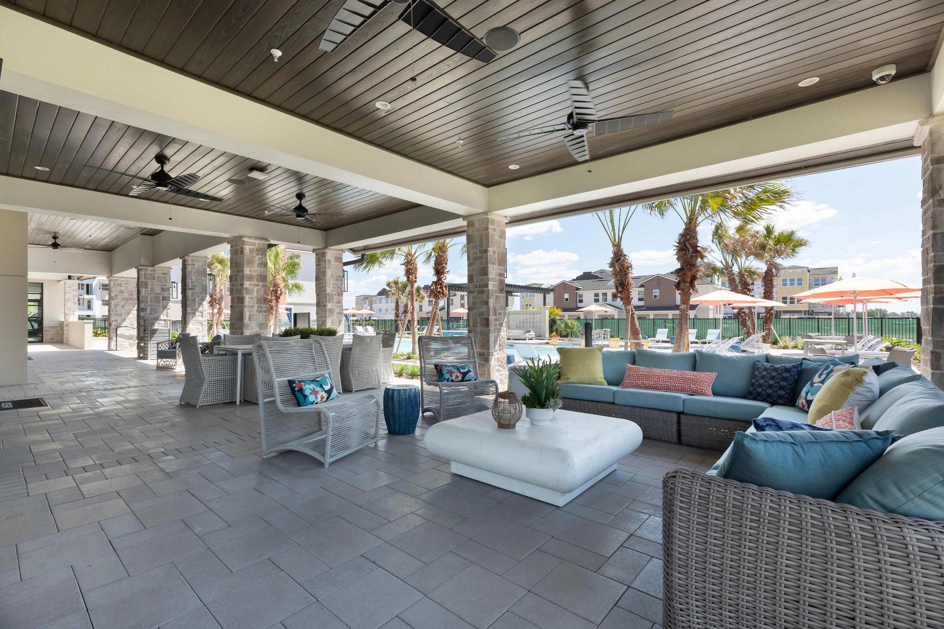 Covered outdoor lounge area with wicker furniture, cushions, a white table, ceiling fans, and a view of a pool and palm trees in the background.
