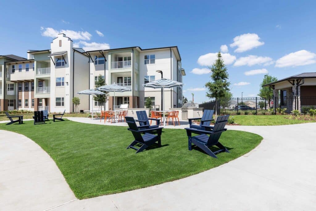 Outdoor seating area with blue chairs on green lawn, tables with striped umbrellas, and a modern apartment building in the background under a blue sky.