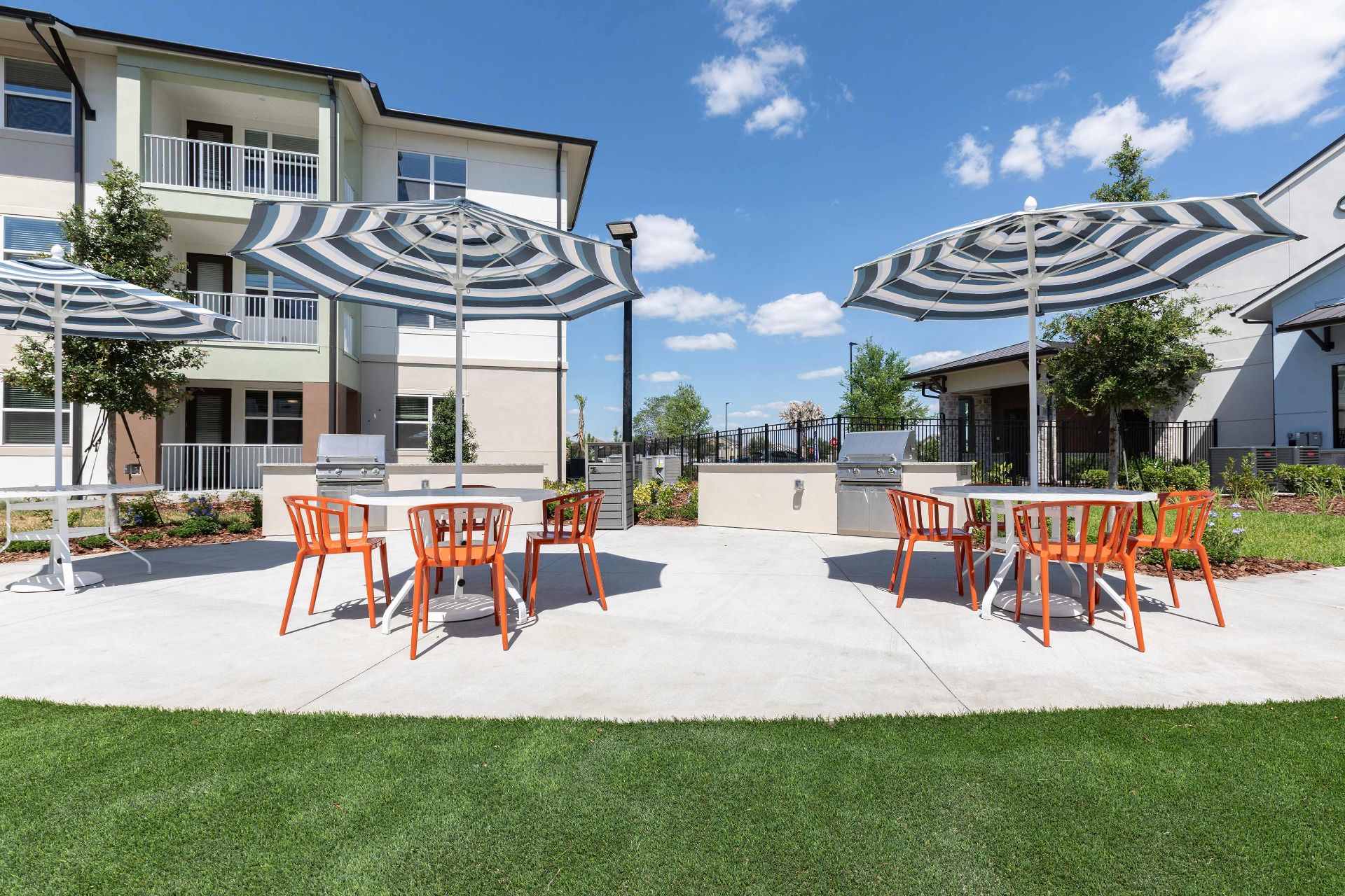 Outdoor patio area with tables, orange chairs, and striped umbrellas on a concrete surface, surrounded by grass and apartment buildings under a blue sky.