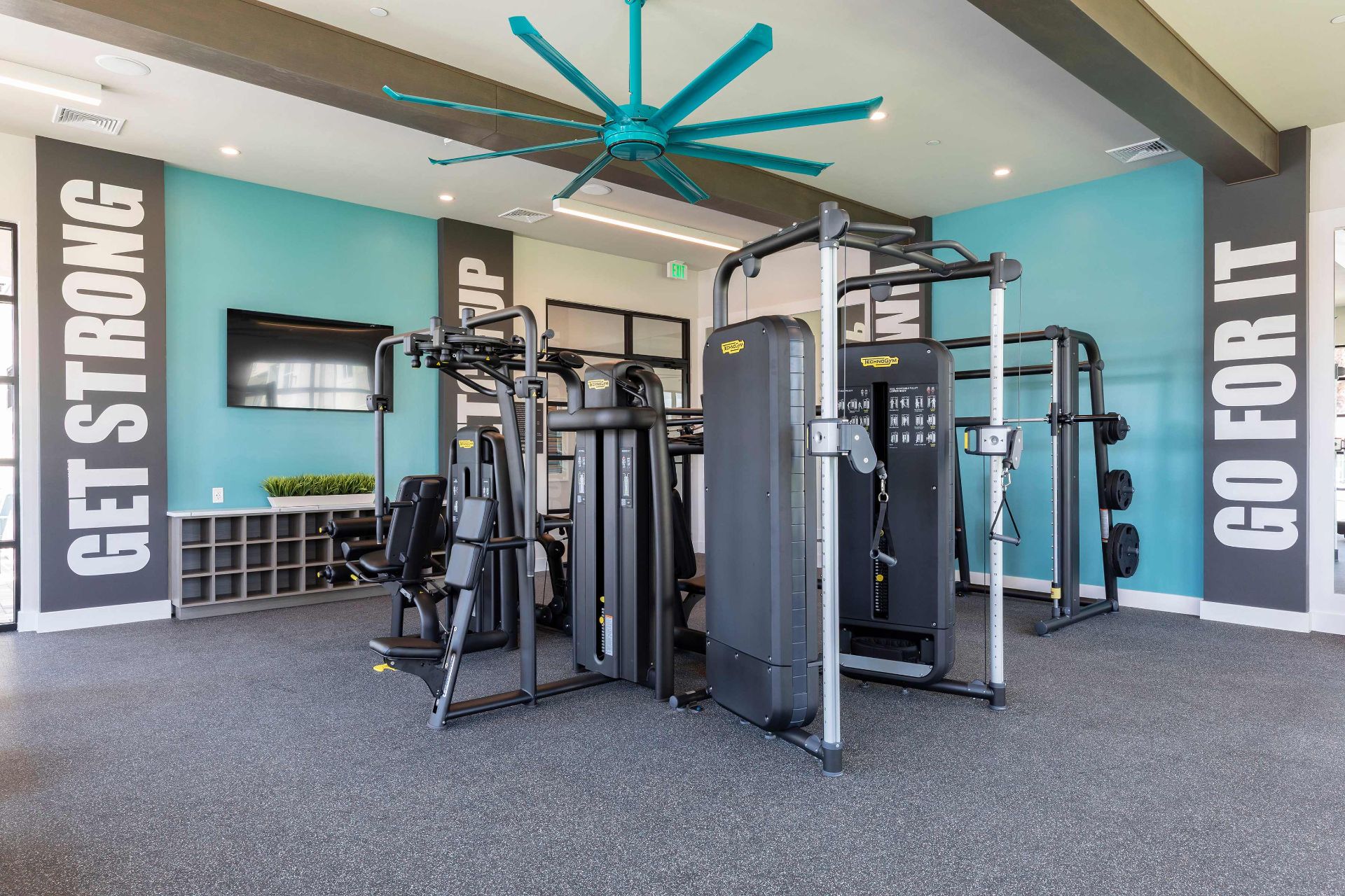 Modern gym with strength training machines, teal accent wall, large ceiling fan, wall-mounted TV, and motivational phrases "GET STRONG" and "GO FOR IT" on the walls.