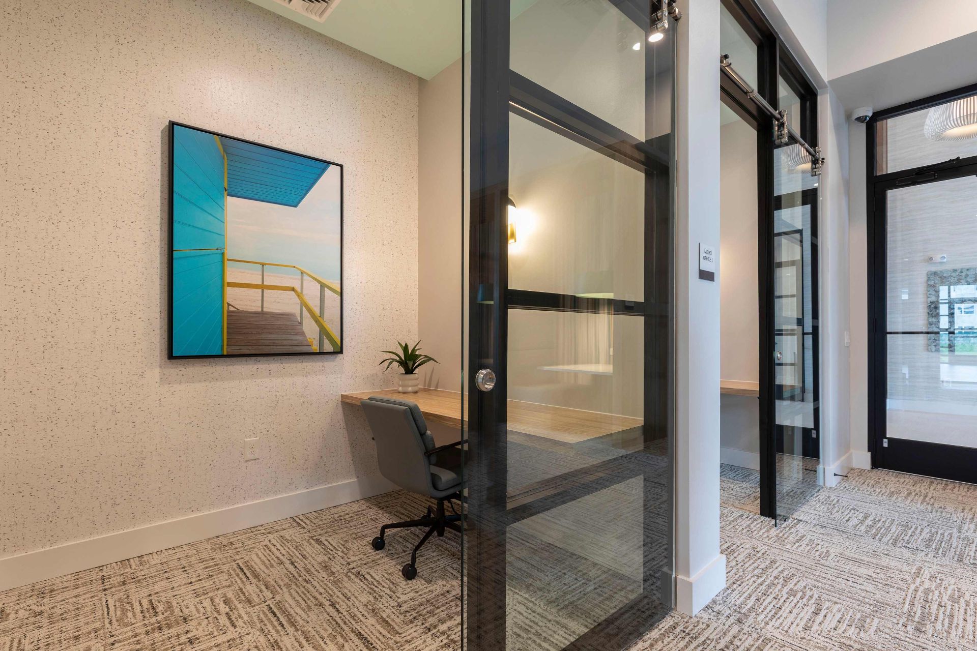 Modern office with a glass door, a desk and chair, a small potted plant, and a wall painting of a boardwalk by the sea.