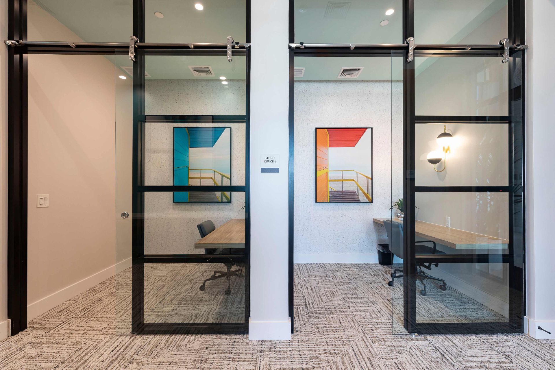 Two modern glass-walled office rooms with desks and chairs; each room features a large framed print of a staircase on the back wall.