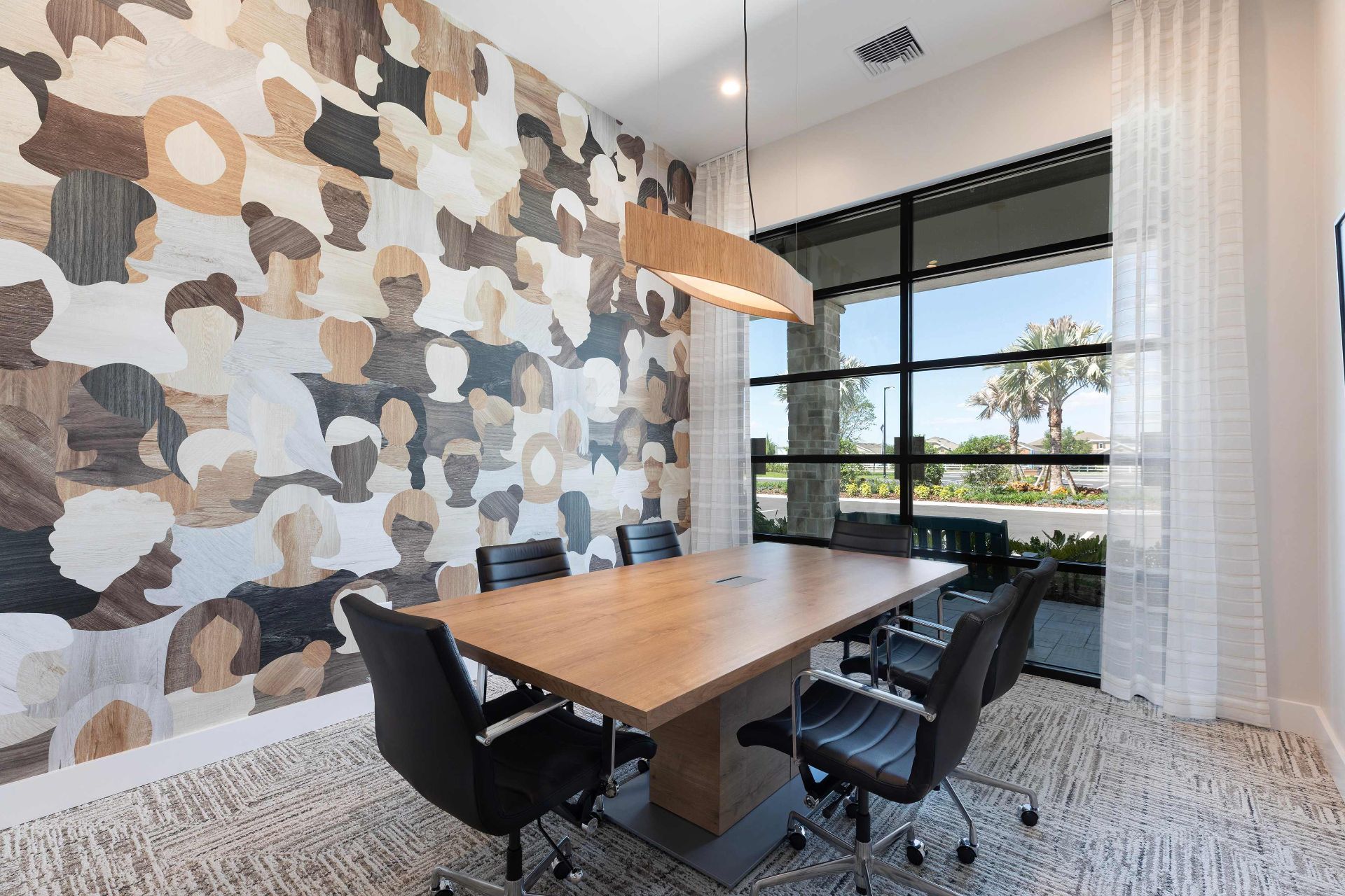 Modern conference room with a wooden table, four black chairs, abstract-patterned accent wall, large window, and natural light.