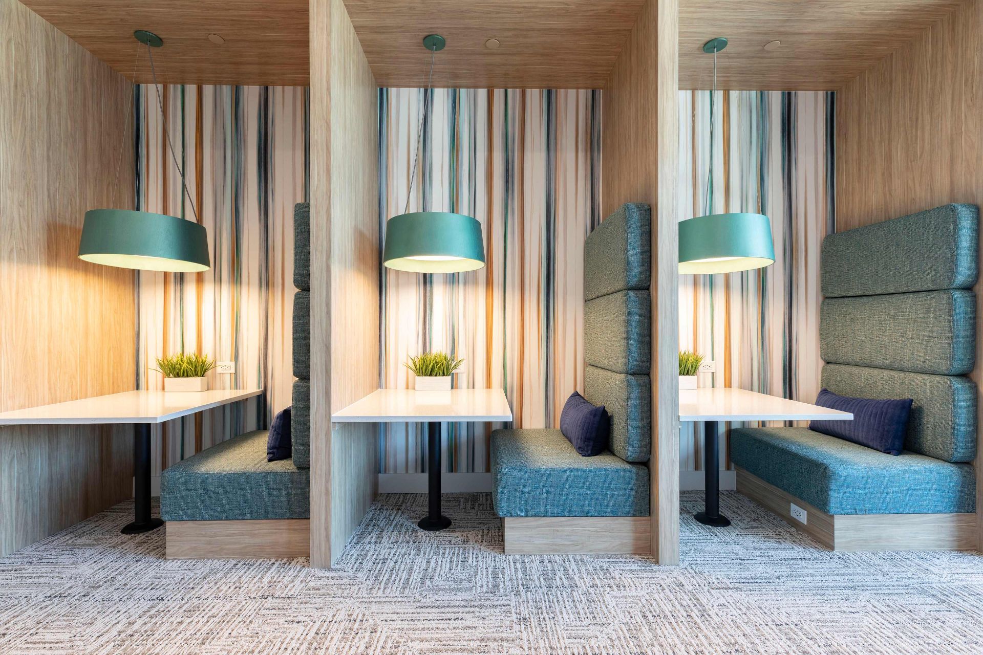 Three modern booth seating areas with light wood panels, blue upholstered benches, white tables, and green pendant lights, each with a small potted plant on the table.
