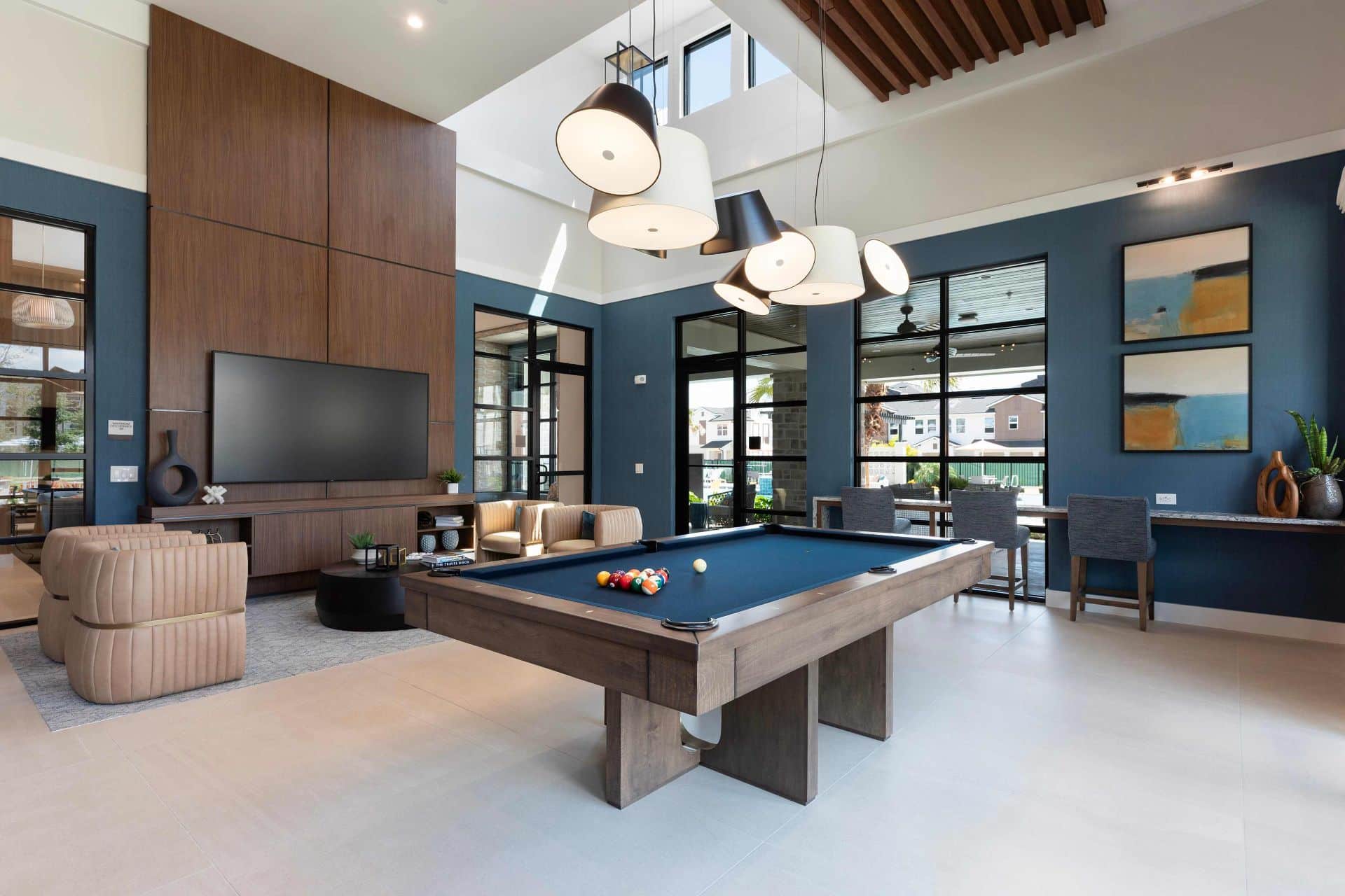 Modern lounge area with a pool table, seating, wall-mounted TV, large windows, contemporary lighting, and artwork on blue walls.
