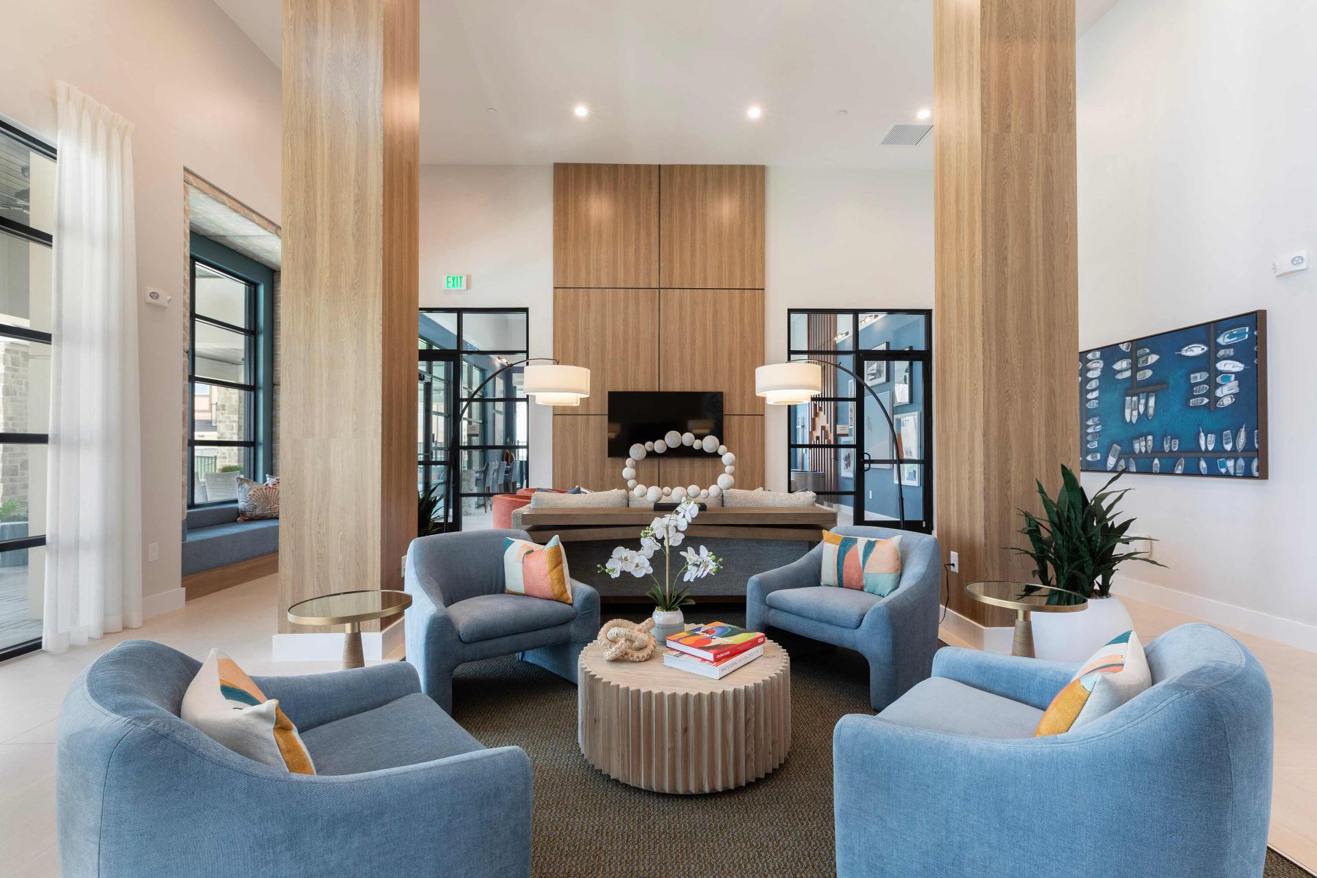 Modern lounge area with four blue armchairs around a round coffee table, decorative plants, books, and large floor lamps, set against wooden paneling and high ceilings.