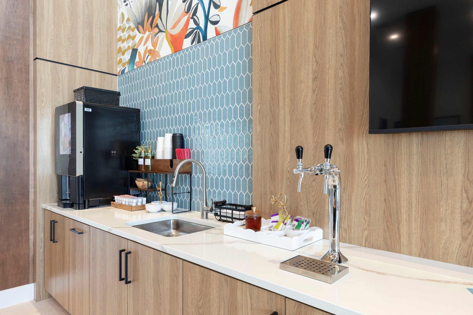 Modern kitchenette with a sink, coffee machine, beverage taps, cups, condiments, and a blue hexagonal tile backsplash against wood-paneled cabinets.