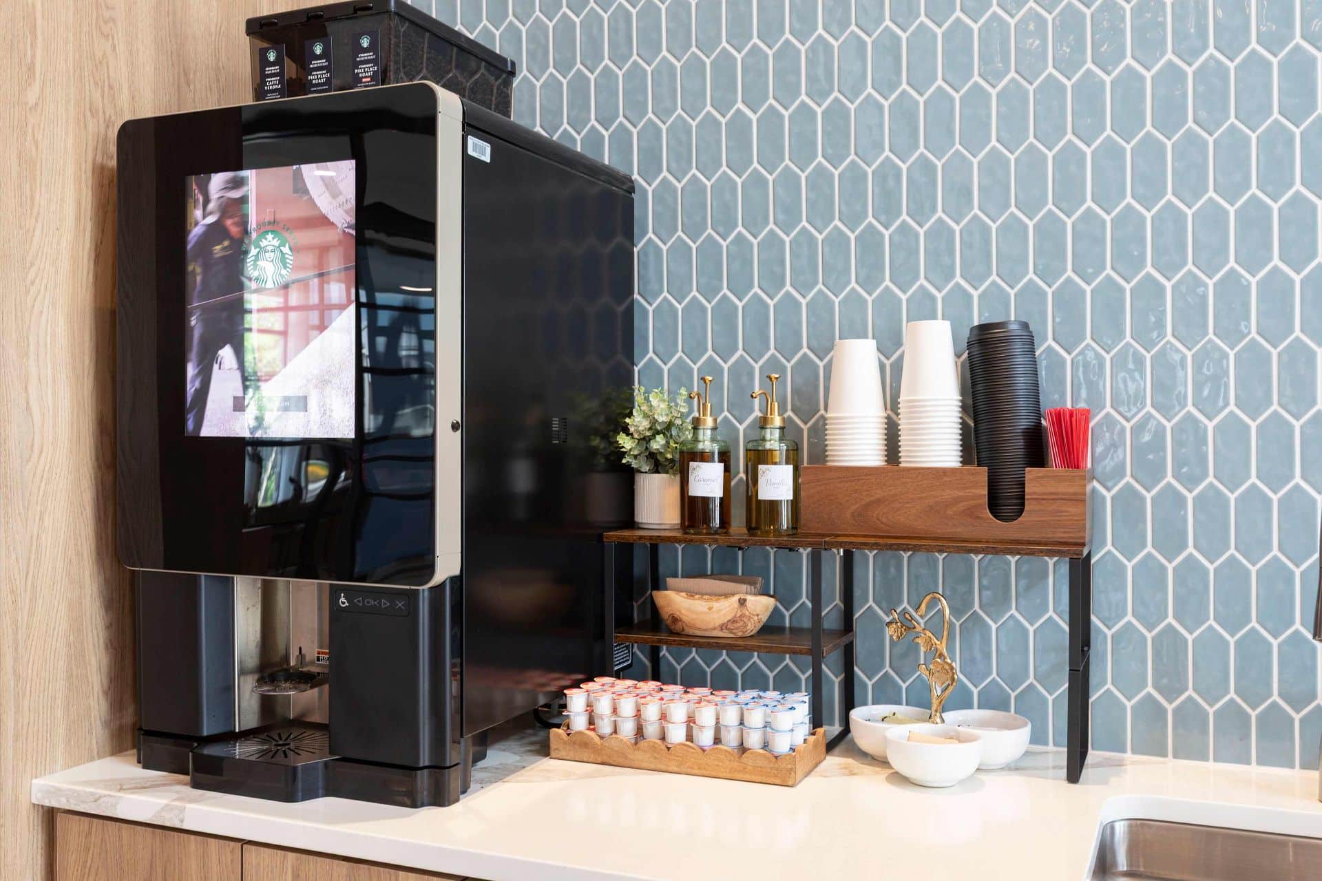 A modern coffee machine sits on a counter next to stacked cups, coffee syrups, stirrers, creamers, and a small decorative plant against a blue tiled wall.