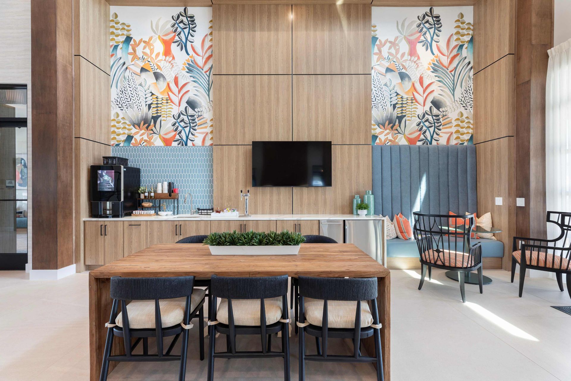 Modern lounge area with a wooden dining table, black chairs, wall-mounted TV, beverage station, plants, and colorful botanical wall art.