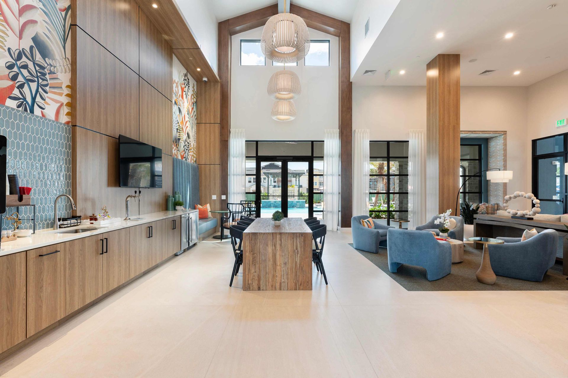 Modern communal lounge with high ceilings, wood accents, a long table, blue chairs, large windows, and decorative lighting fixtures.