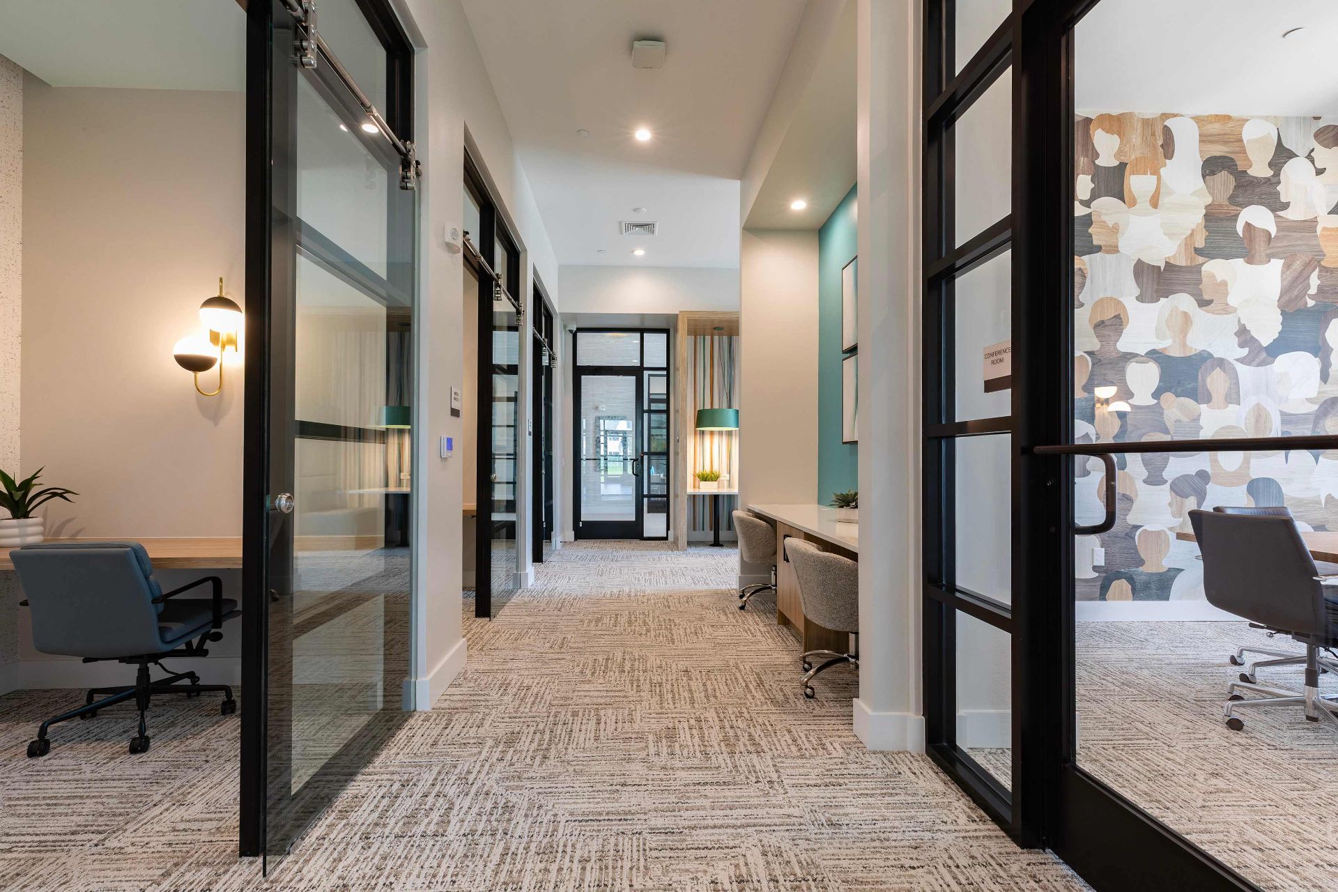 Modern office hallway with glass-walled rooms, carpeted floor, neutral decor, and a feature wall with abstract art in one room. Multiple desks and chairs are visible throughout.