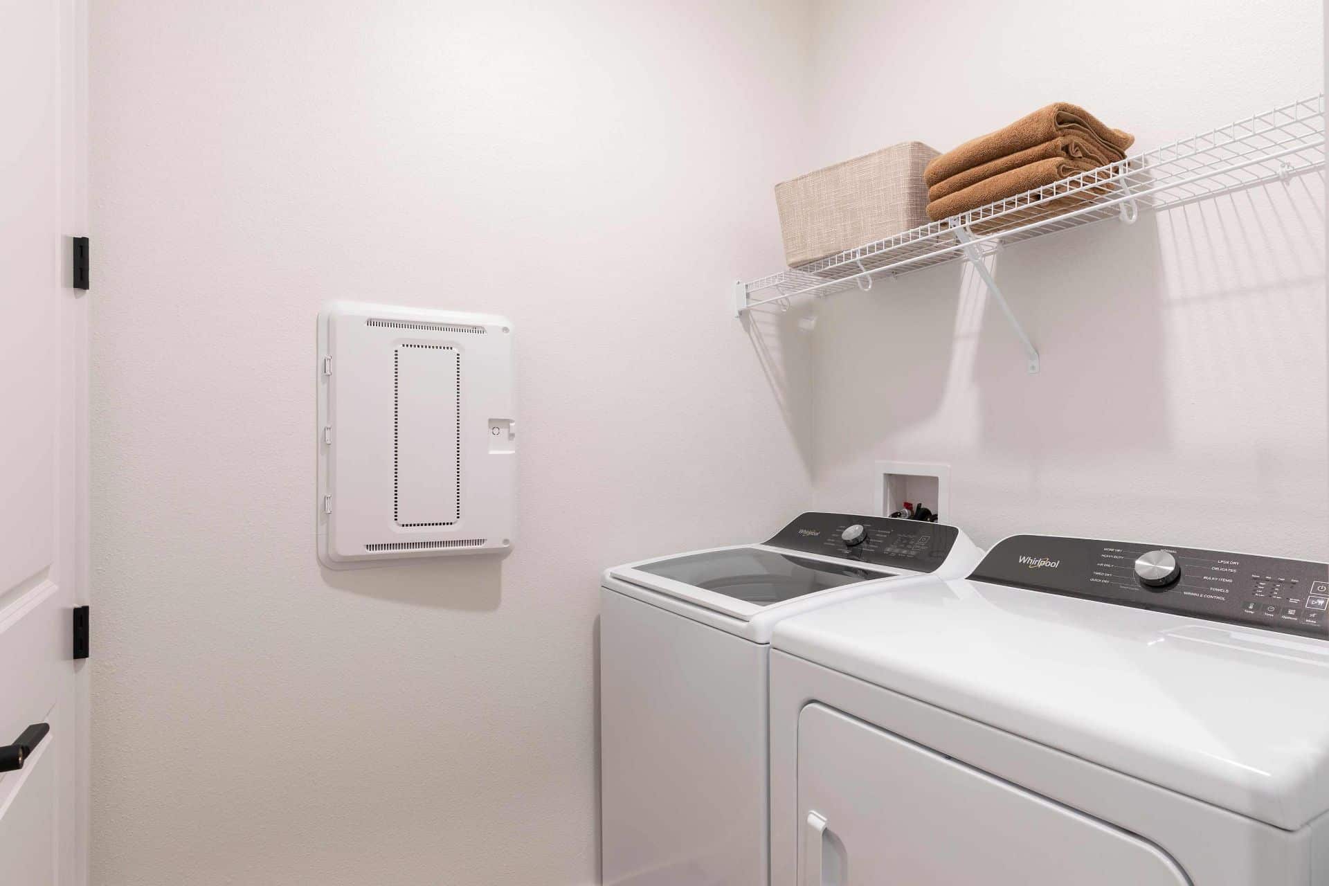 Laundry room with a washing machine and dryer side by side, a wire shelf above holding a basket and folded towels, and an electrical panel on the wall.