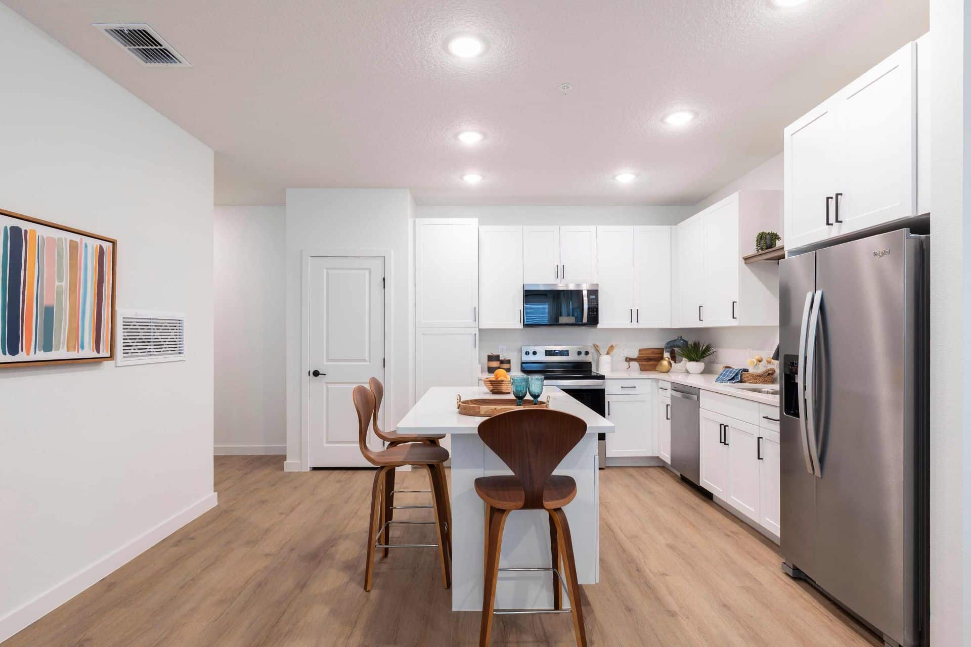 Modern kitchen with white cabinets, stainless steel appliances, an island with two wooden chairs, and light wood flooring under recessed ceiling lights.