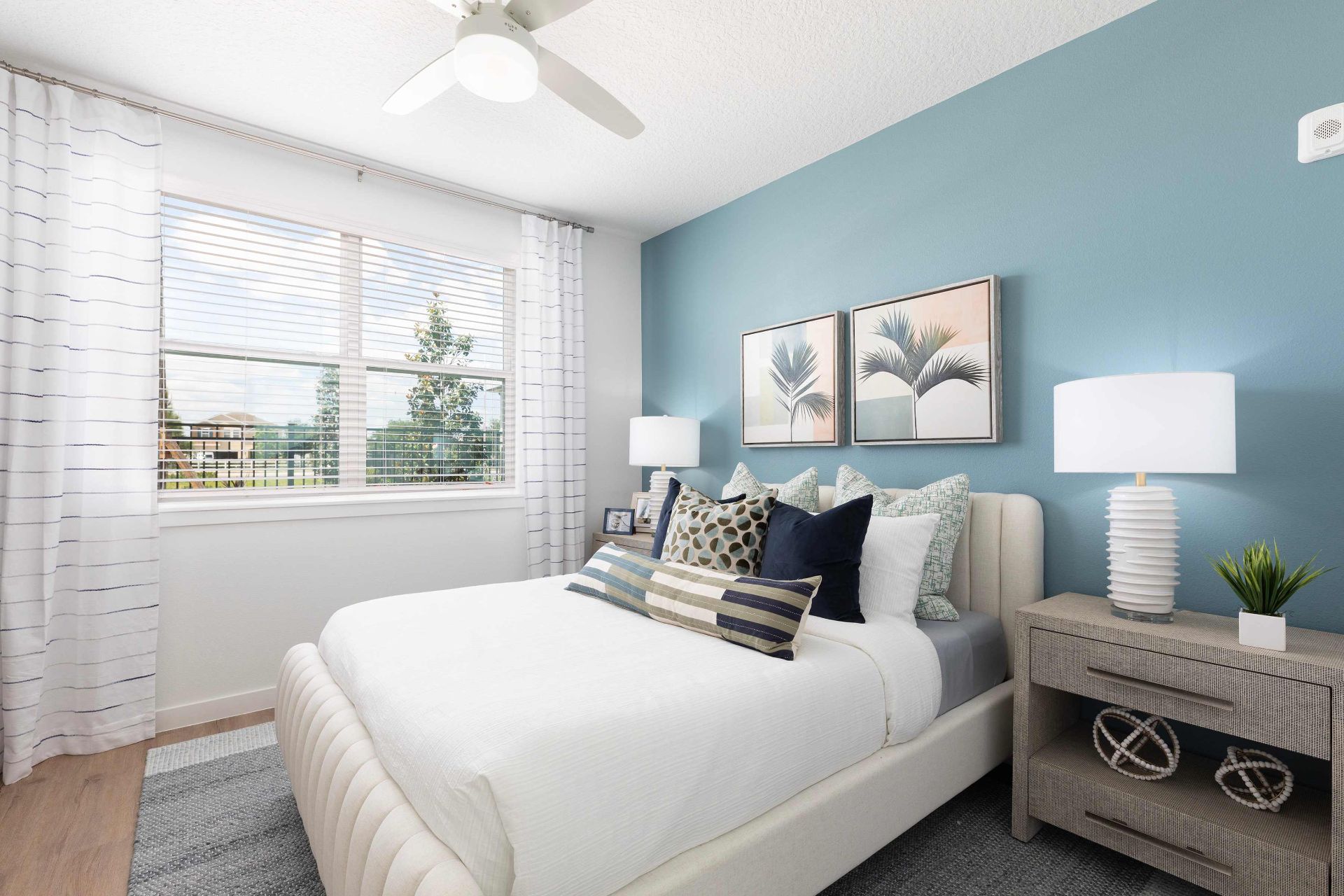 A modern bedroom with a blue accent wall, white bed, two bedside tables with lamps, decorative pillows, framed art, and a window with white curtains.