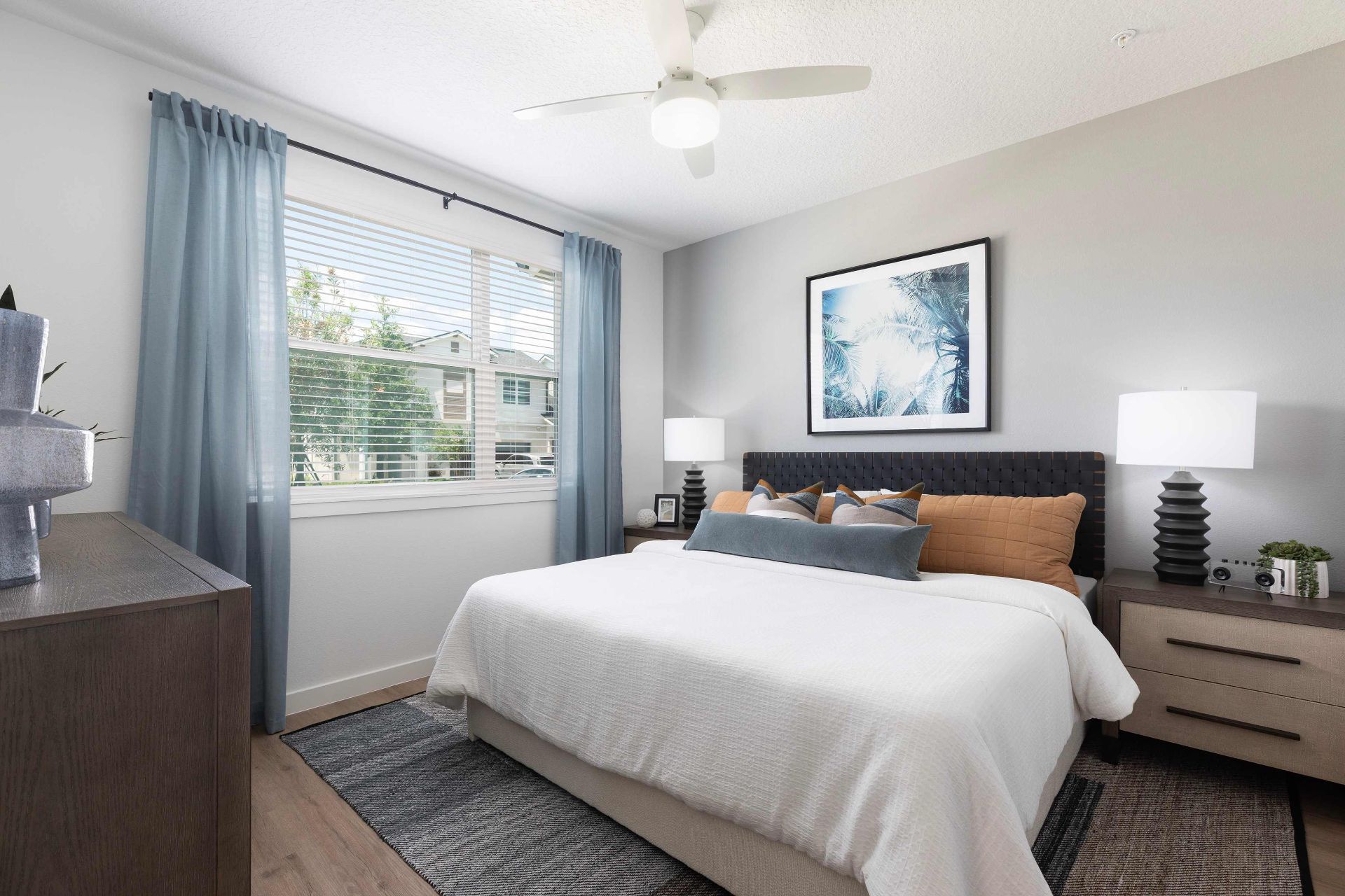 A modern bedroom with a neatly made bed, two bedside tables with lamps, a dresser, light blue curtains, and a ceiling fan.