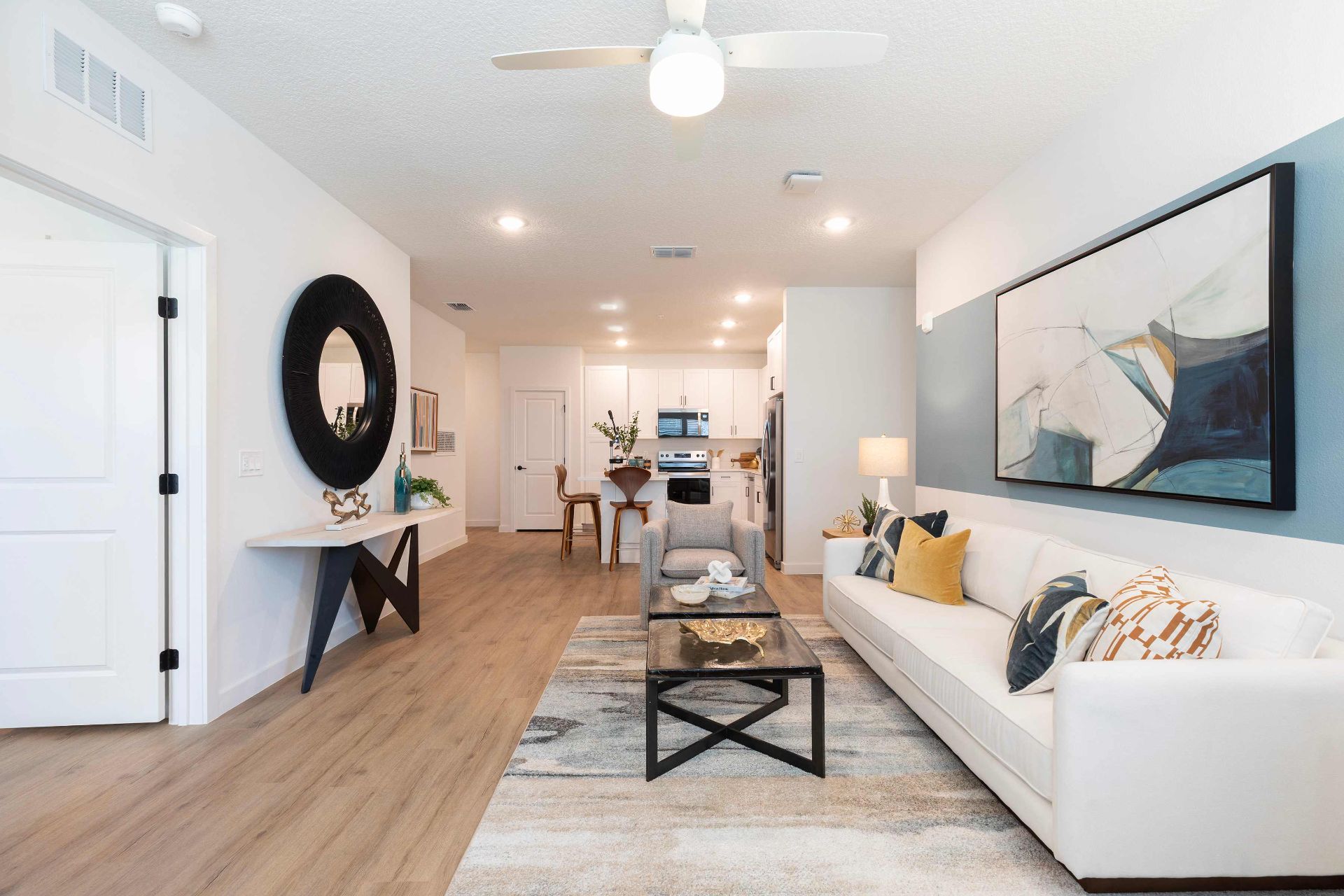 Modern living room with white sofa, abstract art, round mirror, and wooden floors, leading to a kitchen and dining area in the background.