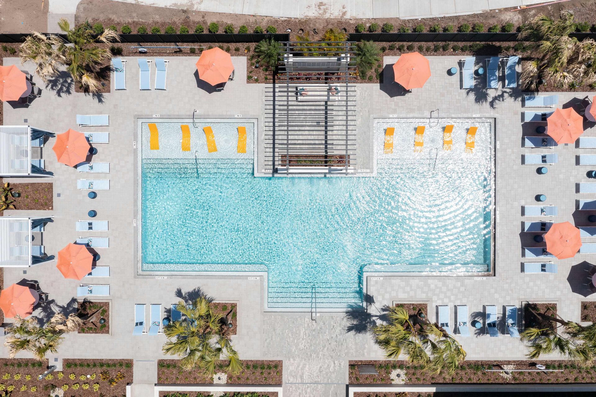 Aerial view of a rectangular swimming pool surrounded by lounge chairs, orange umbrellas, cabanas, and landscaped greenery.