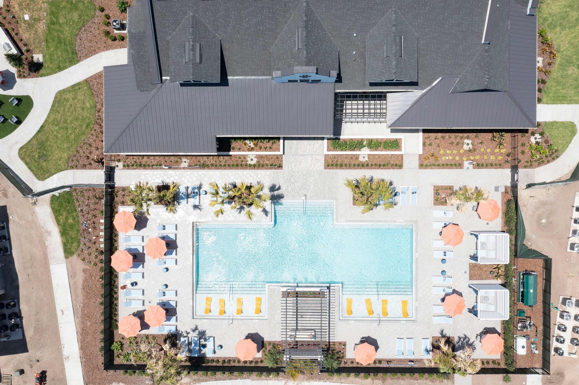 Aerial view of a rectangular swimming pool with lounge chairs, umbrellas, palm trees, and a building at the top of the image.