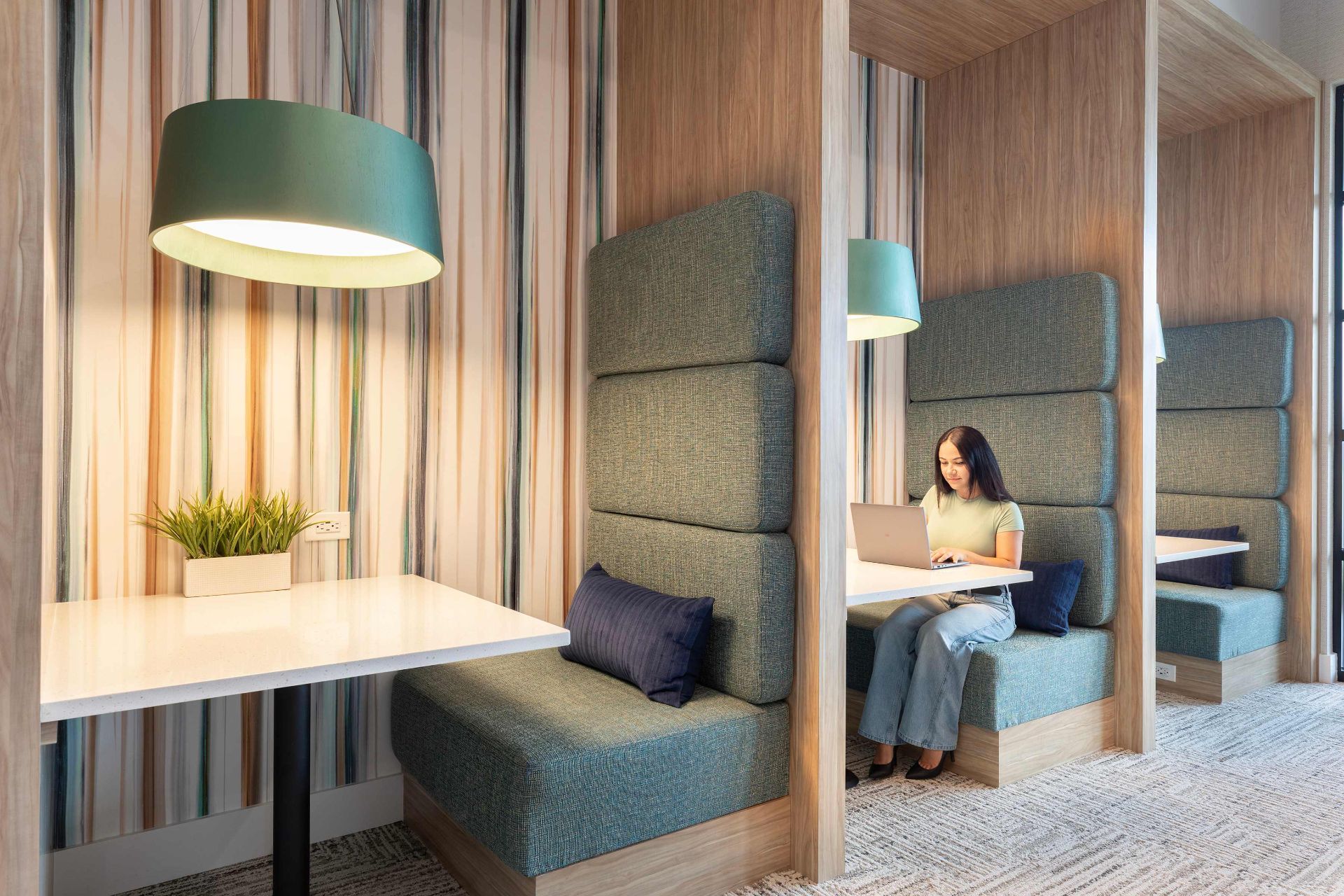 Woman sitting and working on a laptop in a modern booth seating area with padded benches, tables, pendant lights, and striped wallpaper.