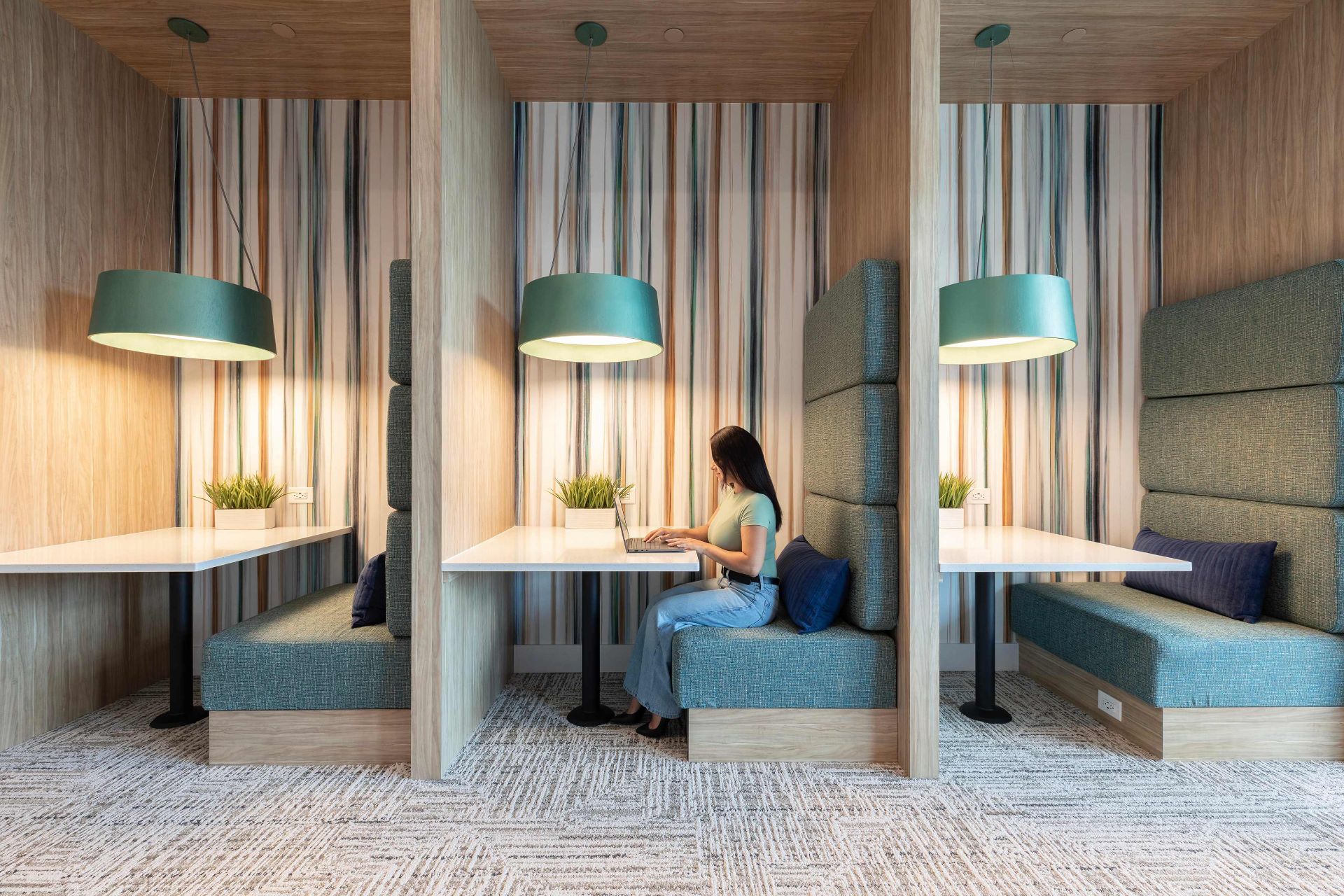 A woman sits alone in a modern booth workspace with a laptop, surrounded by wood and teal furnishings and striped wall panels.