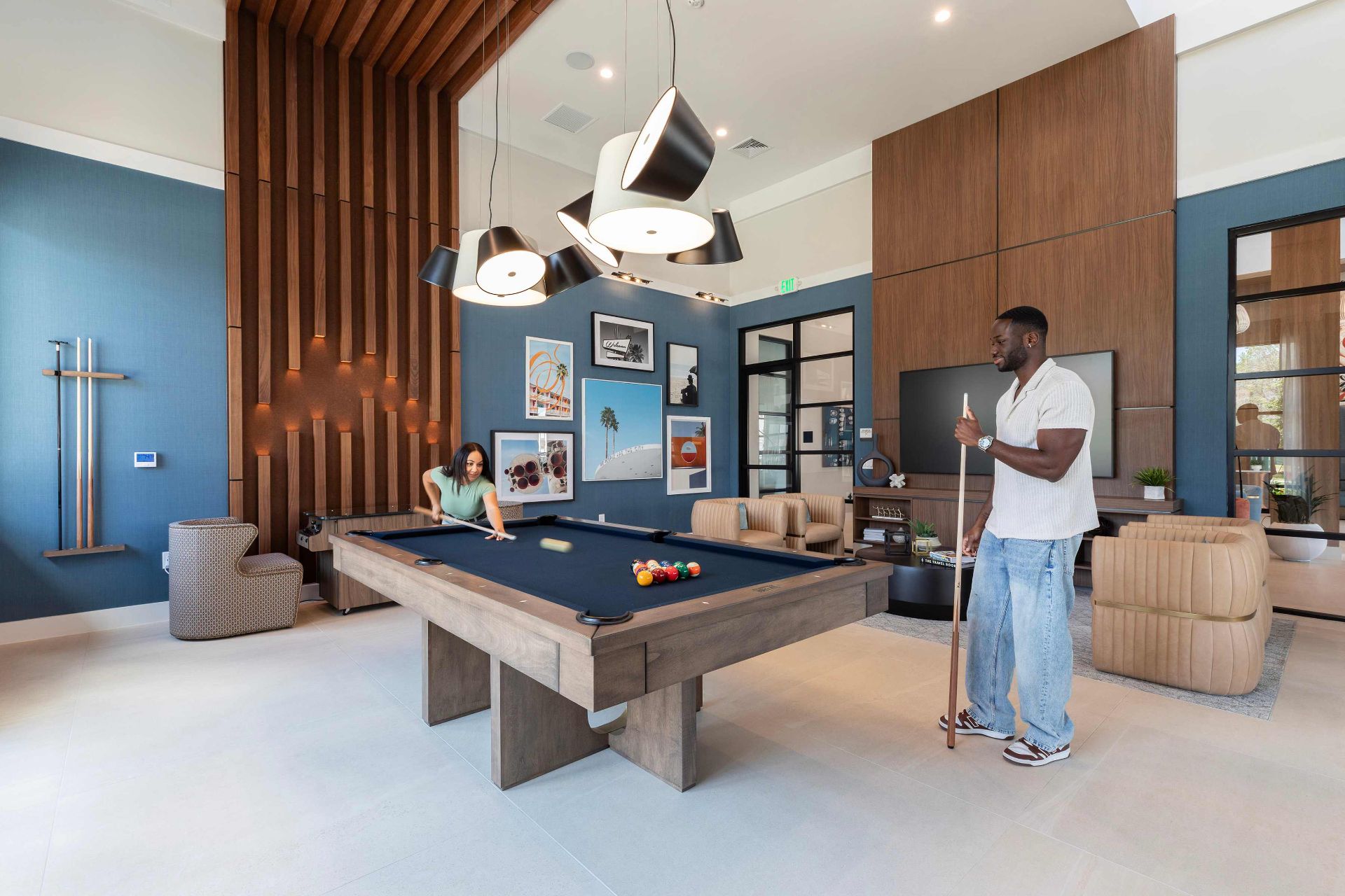Two people play pool in a modern, well-lit lounge with contemporary furniture, wall art, and decorative lighting fixtures.