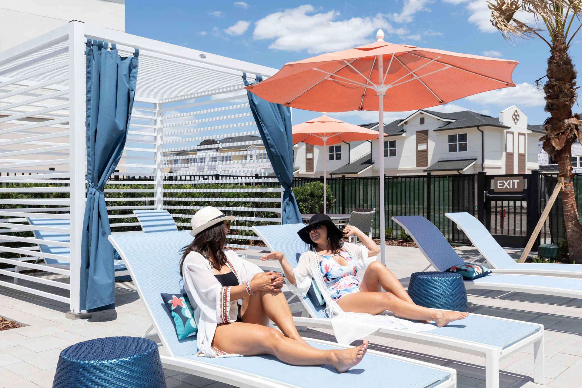 Two women in sun hats relax on lounge chairs by a pool under an orange umbrella, with modern apartments and a cabana in the background.