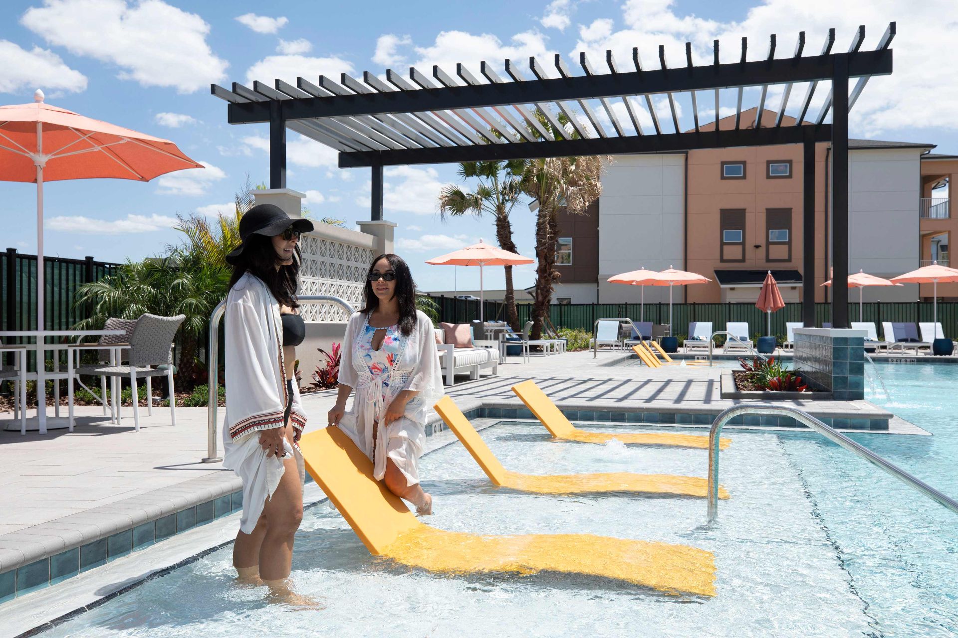 Two women wearing sunglasses and cover-ups stand and sit on yellow loungers in a shallow pool area, with poolside furniture and umbrellas in the background.
