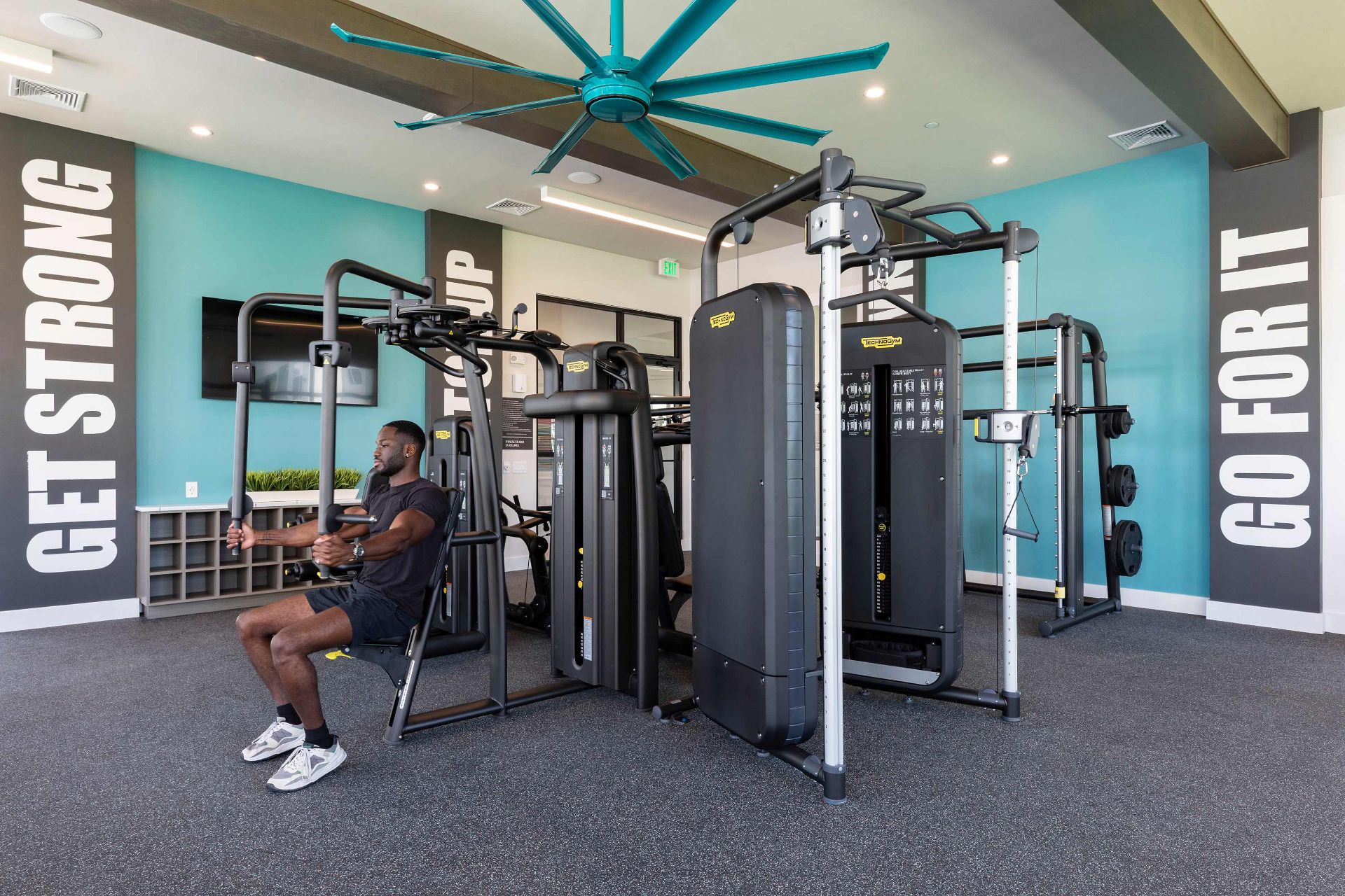 A man uses a seated weight machine in a modern gym with large equipment, teal walls, motivational signs, and a ceiling fan.