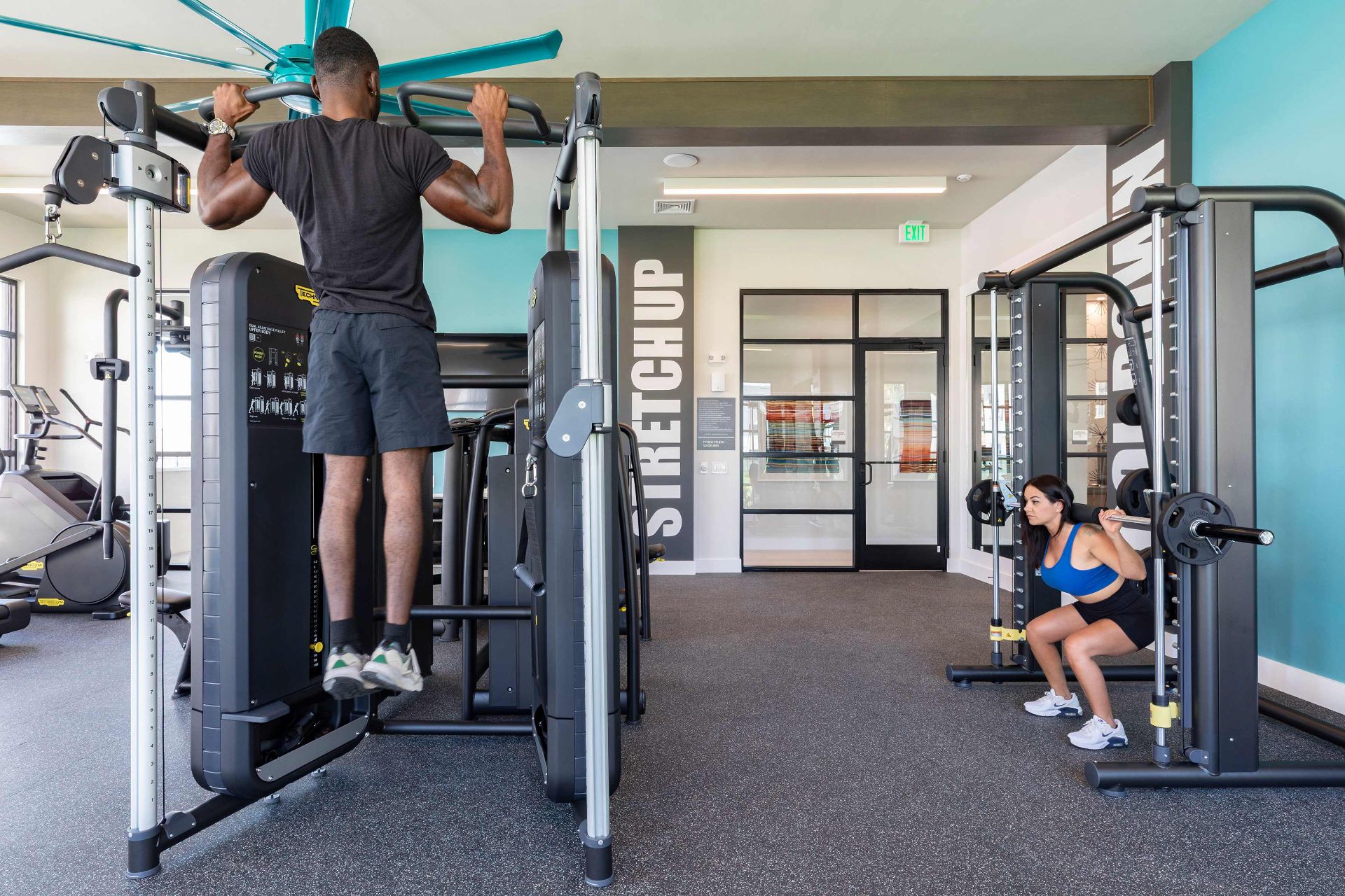 A man performs pull-ups on a gym machine while a woman does squats with a barbell in a modern fitness center.