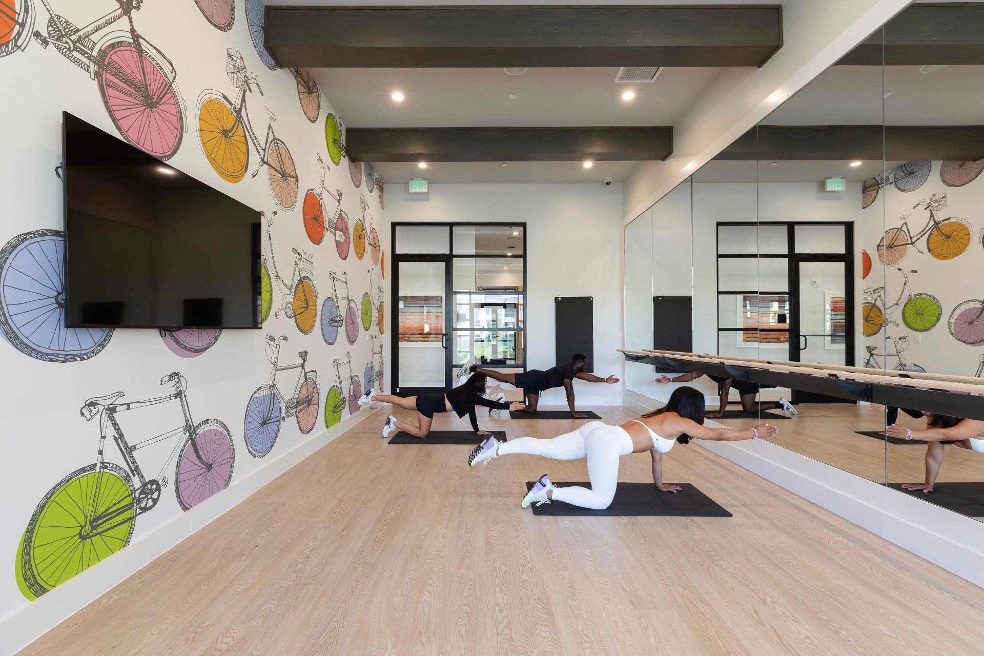 Two people exercise on mats in a brightly lit fitness room with a wall mural of colorful bicycles and a large mirror reflecting the space.